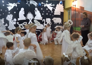 Na tle bożonarodzeniowej dekoracji tańczą w czteroosobowych zespołach dzieci w strojach aniołów.